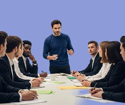 Man in blue sweater leading business meeting with eight colleagues sitting around a table with papers.