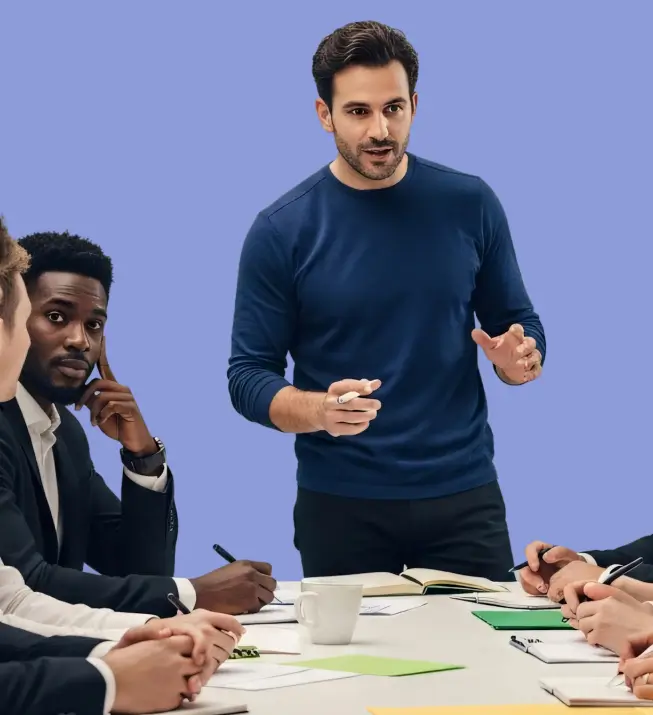 Man in blue sweater standing and speaking to seated colleagues in a business meeting.